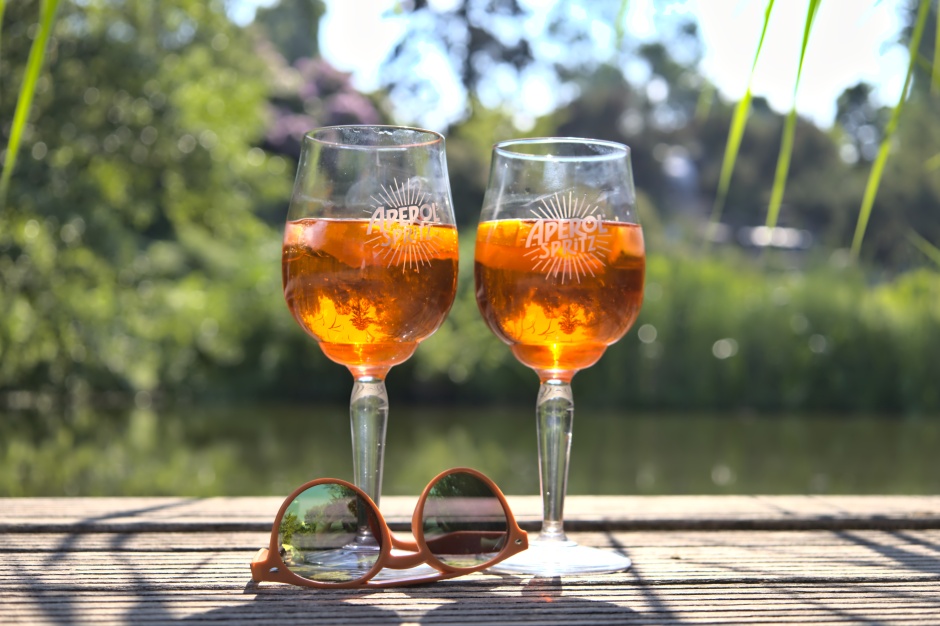 Zwei gefüllte Gläser mit der Aufschrift "Aperol Spritz" stehen auf einem Steg zu einem See. Am Fuße des einen Weinglases liegt eine Sonnenbrille in der sich der See spiegelt.